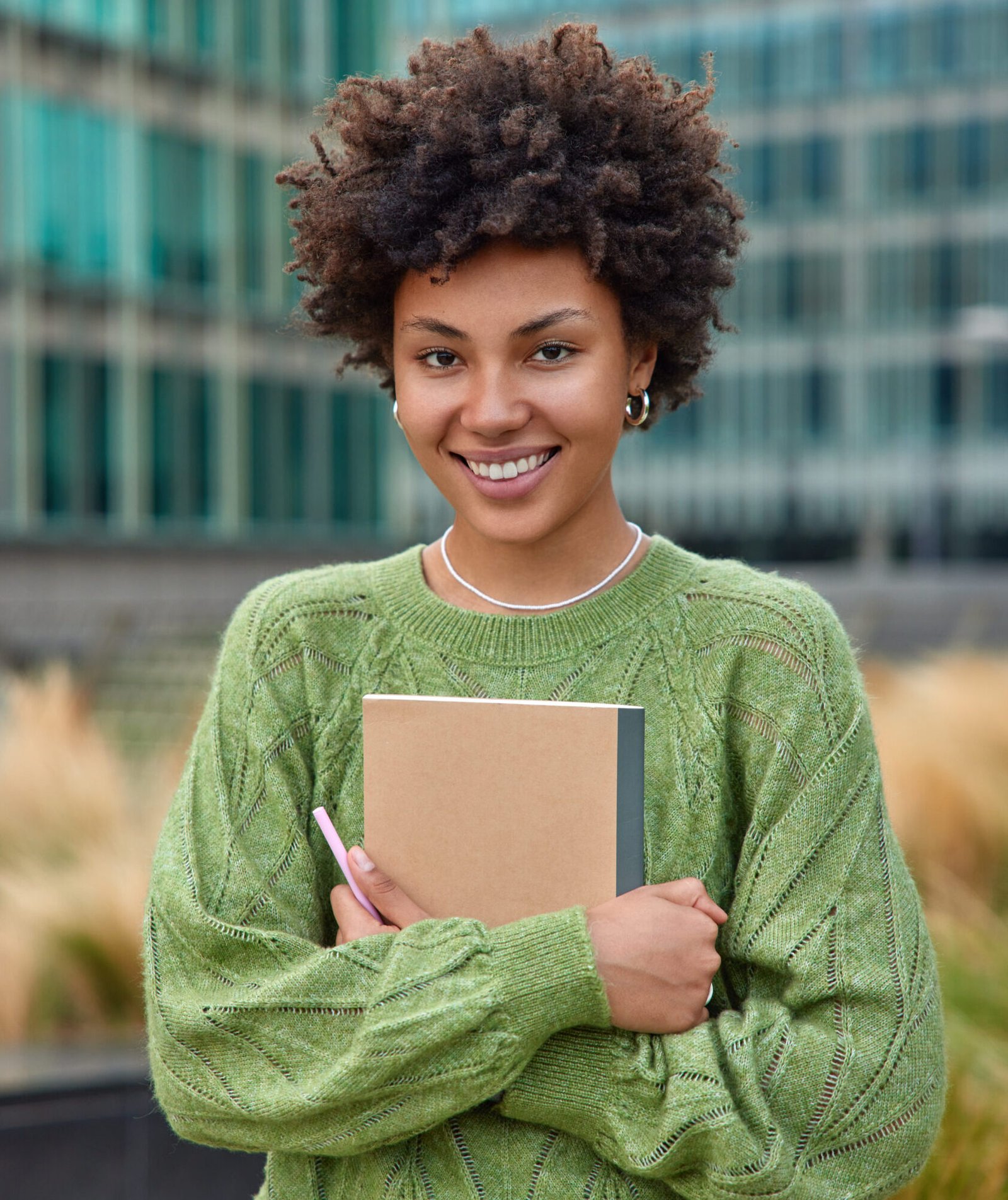 Vertical shot of happy young woman with curly hair holds notepad and pen makes notes what she observes around in city dressed in casual green jumper poses outdoors against blurred background